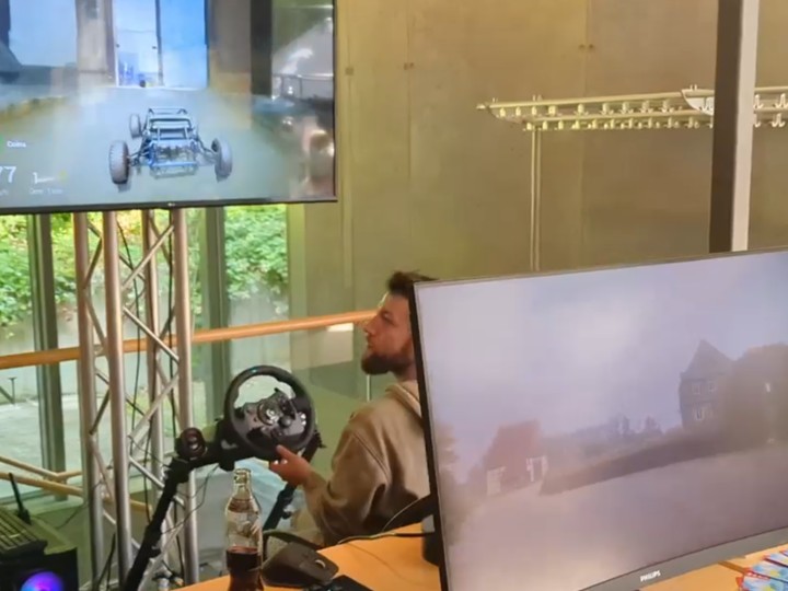 Person using a racing-wheel controller to drive through a photoreal scanned environment on screen, an interactive virtual tour demo.