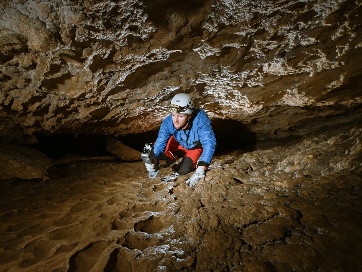 Caver in a helmet crawling through a tight cave passage with a handheld scanner, surveying difficult exploration terrain.