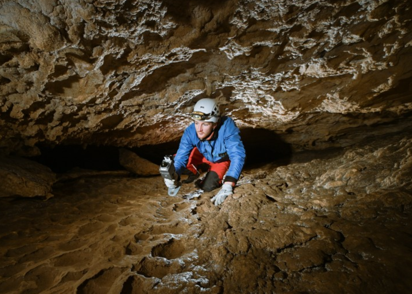 Surveyor in a helmet crawling through a narrow cave with a handheld Lixel scanner, mobile SLAM capture in a GPS-denied environment.